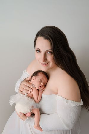 A mother in a white dress cradles her sleeping newborn in an intimate studio portrait series against a neutral background.