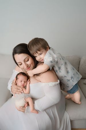 Tender family moment showing mother resting on white couch with newborn and toddler cuddling close together.
