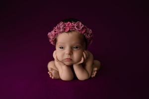 A small baby wearing a delicate pink floral headband rests on a purple backdrop in an artistic newborn portrait.