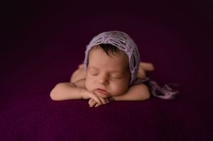 A sleeping newborn wearing a lavender bonnet rests their head on folded arms against a deep purple backdrop.
