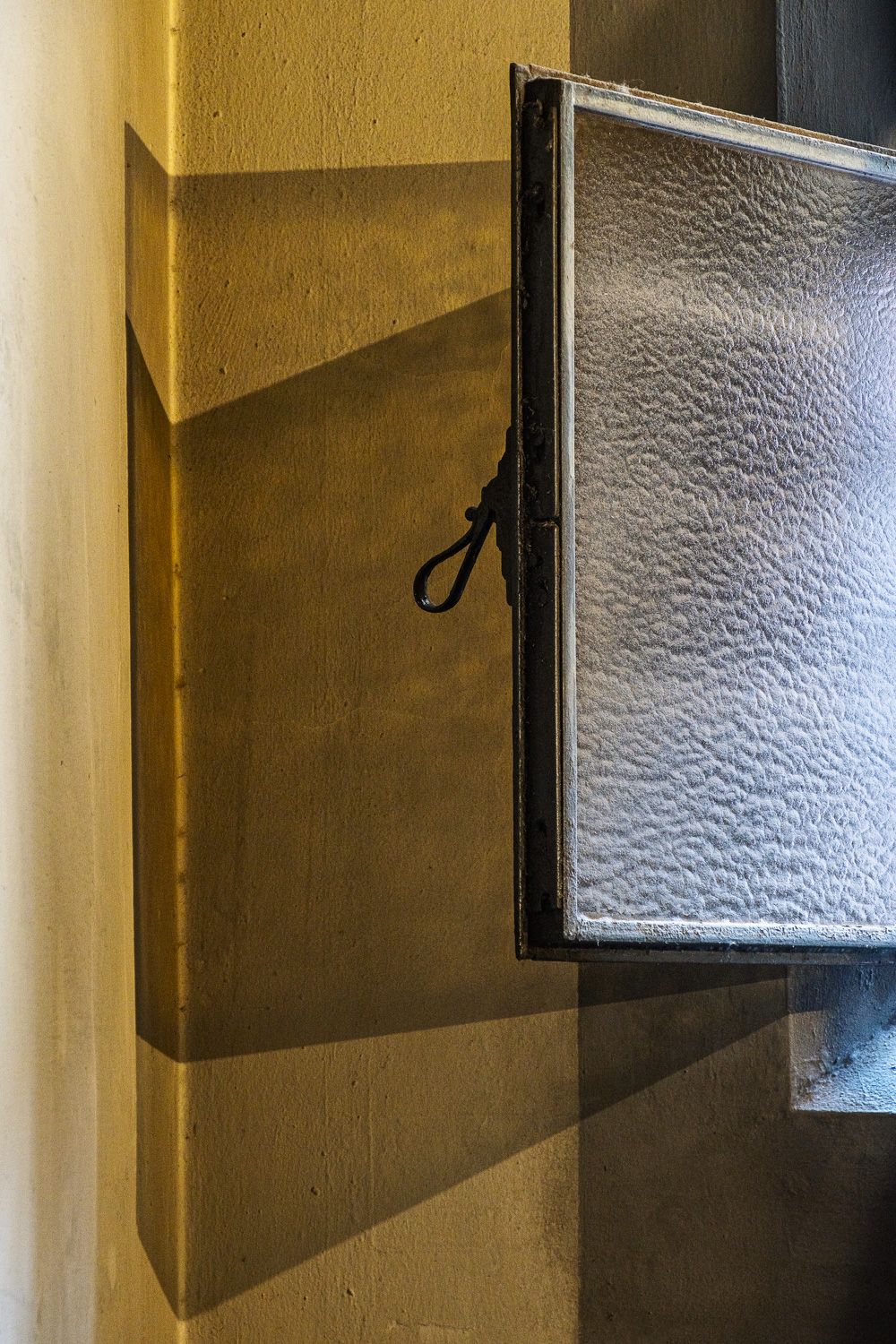 Small frosted glass window panel fitted in black frame casting shadow on white wall in hallway interior.