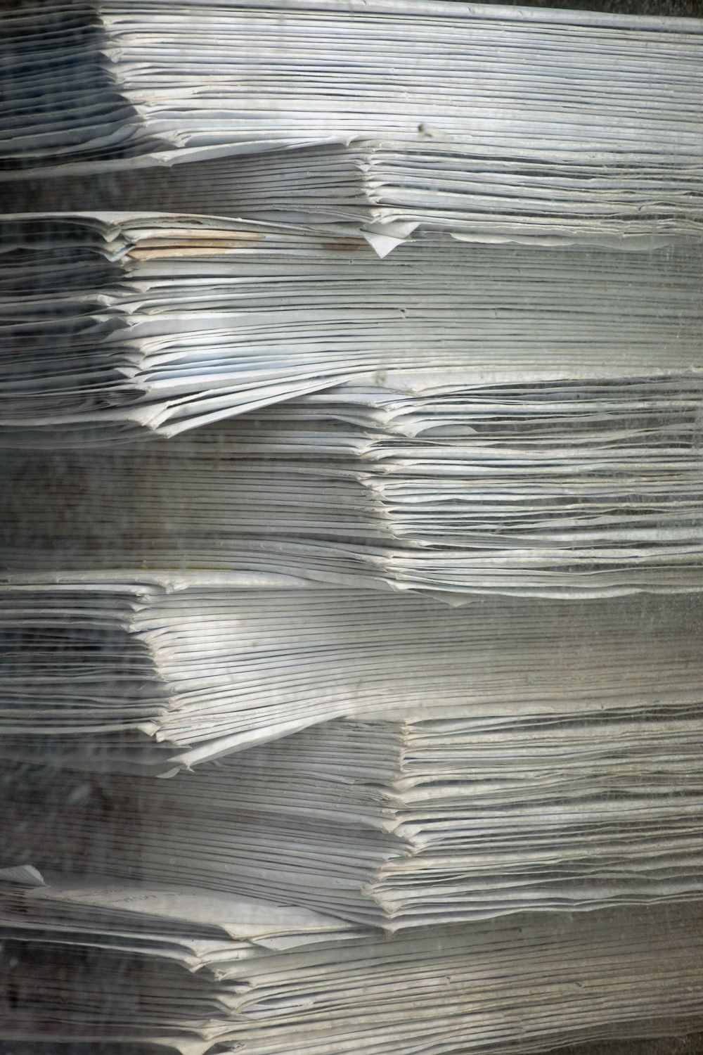 Stacks of old newspapers piled horizontally showing their worn edges in a gray monochromatic pattern.