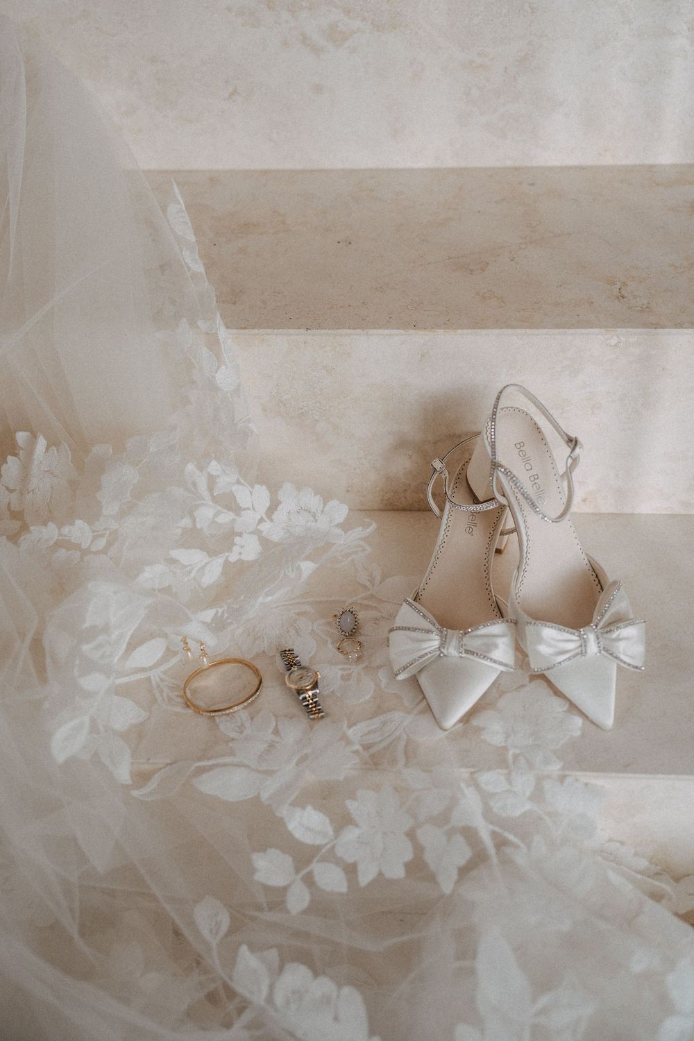 Wedding accessories including white bow heels, jewelry and delicate lace veil laid out on light surface.