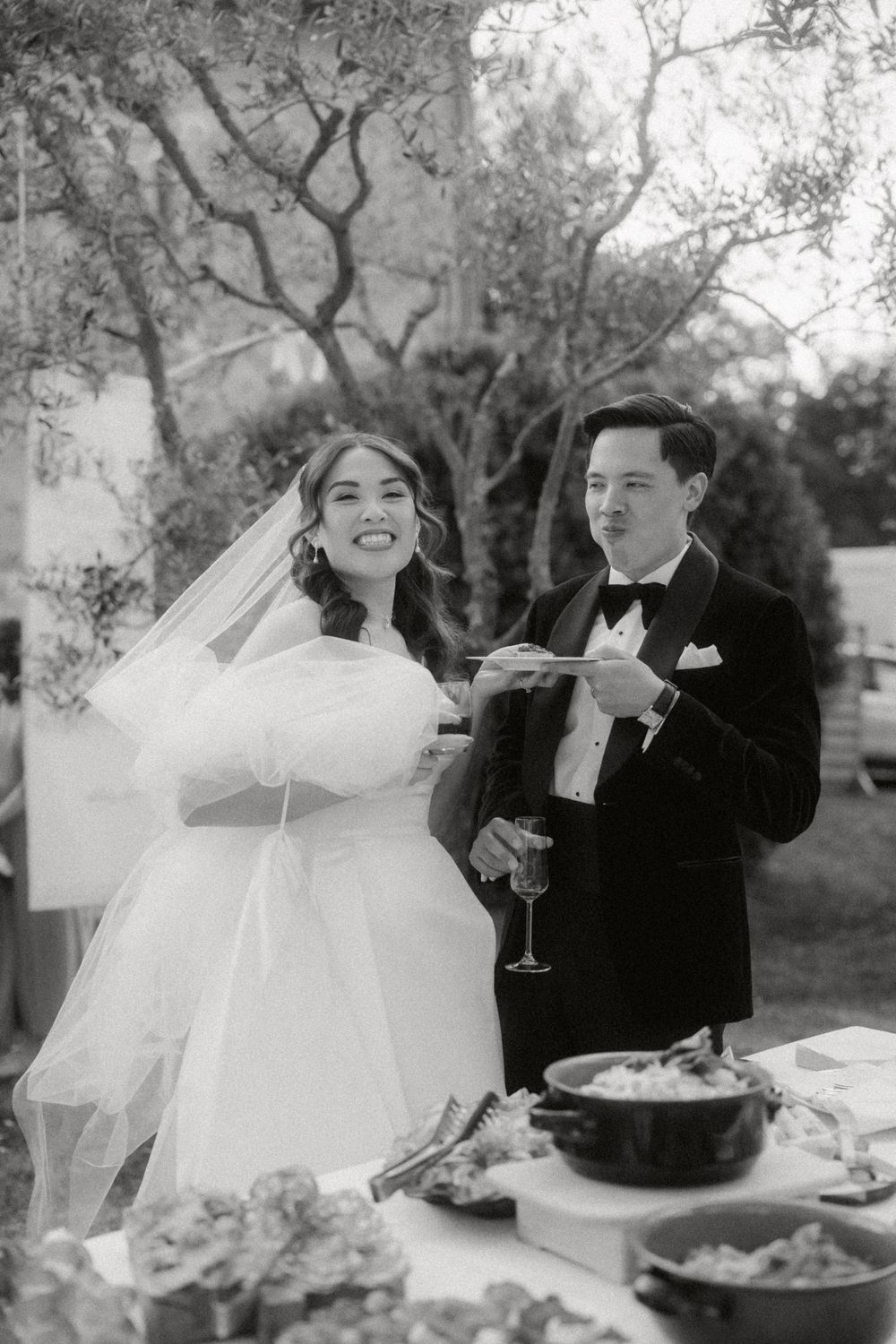 A couple sits together at an outdoor reception table sharing champagne and laughter under trees.