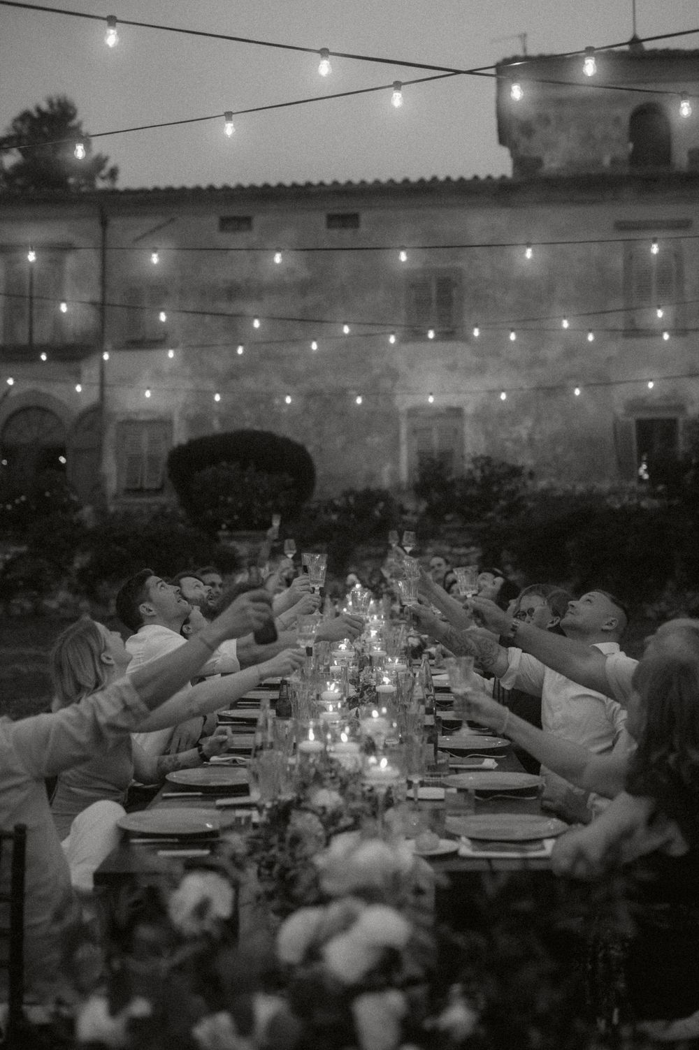 Black and white photo of elegant outdoor dinner party with string lights and long dining table. Tuscany wedding photography