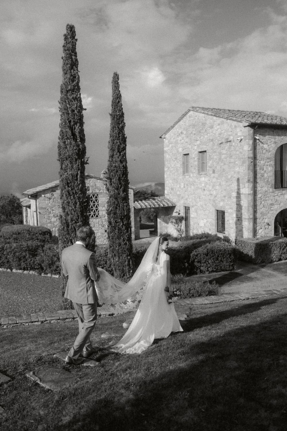 Black and white photo sequence shows couple walking up grassy hillside towards rustic stone villa in Tuscan countryside.