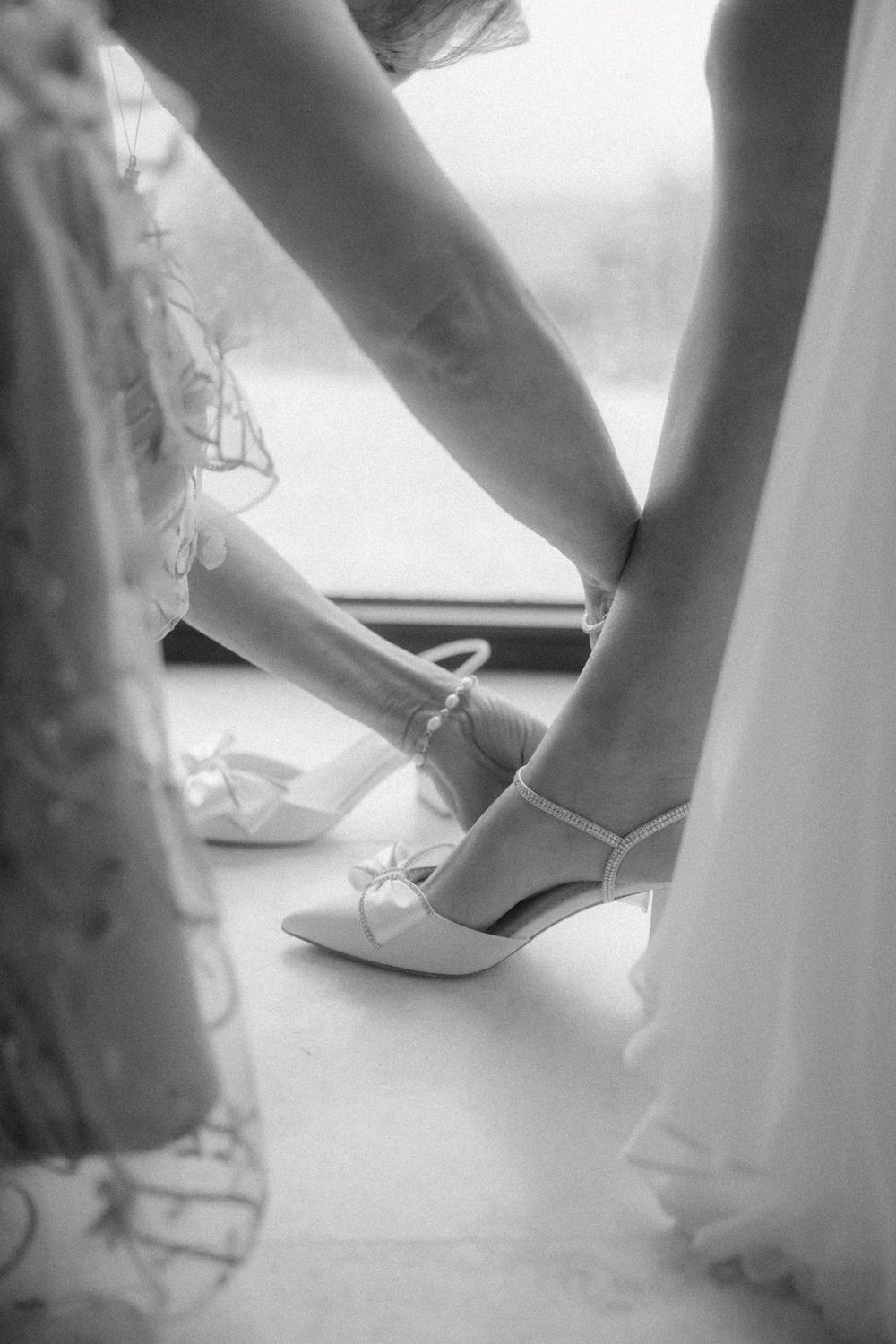 A series of soft-focus black and white photographs showing elegant white bridal shoes being put on during wedding preparation.