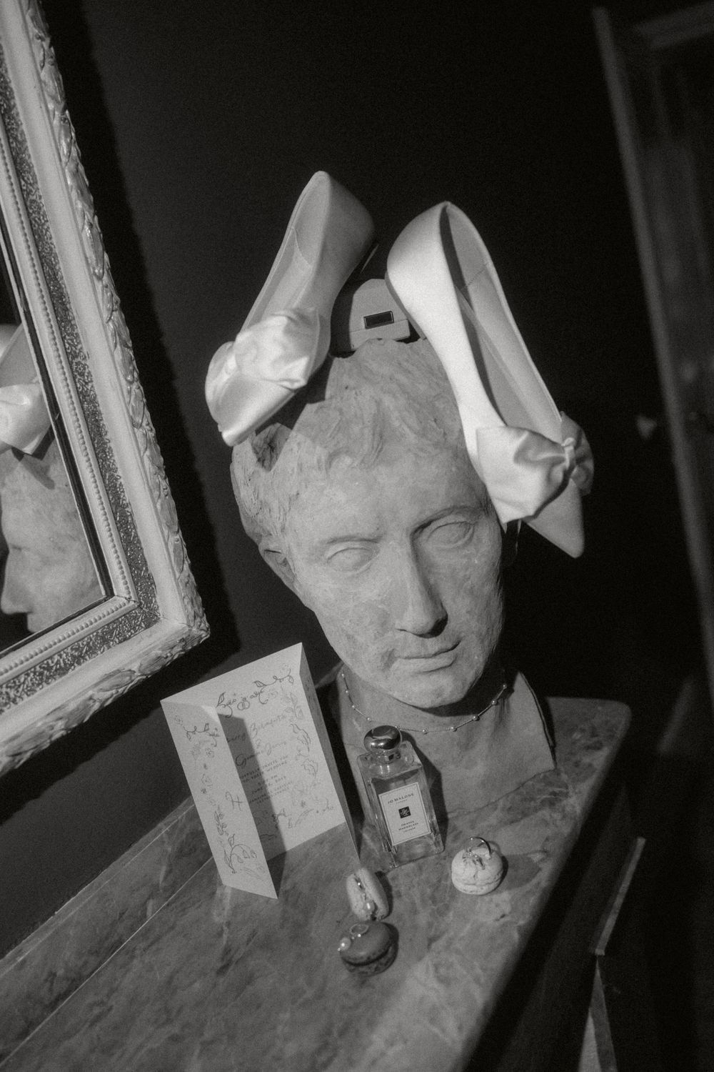 Black and white photograph of a classical bust sculpture with two white shoes balanced on its head next to small decorative items.