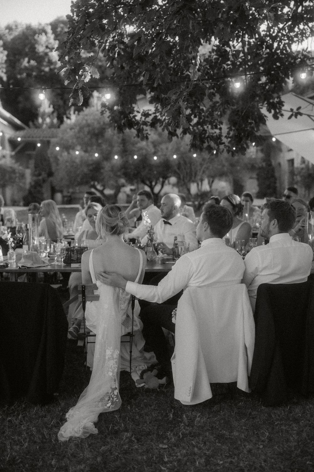 People gathered at an elegant outdoor evening reception under string lights and trees in black and white photography.
