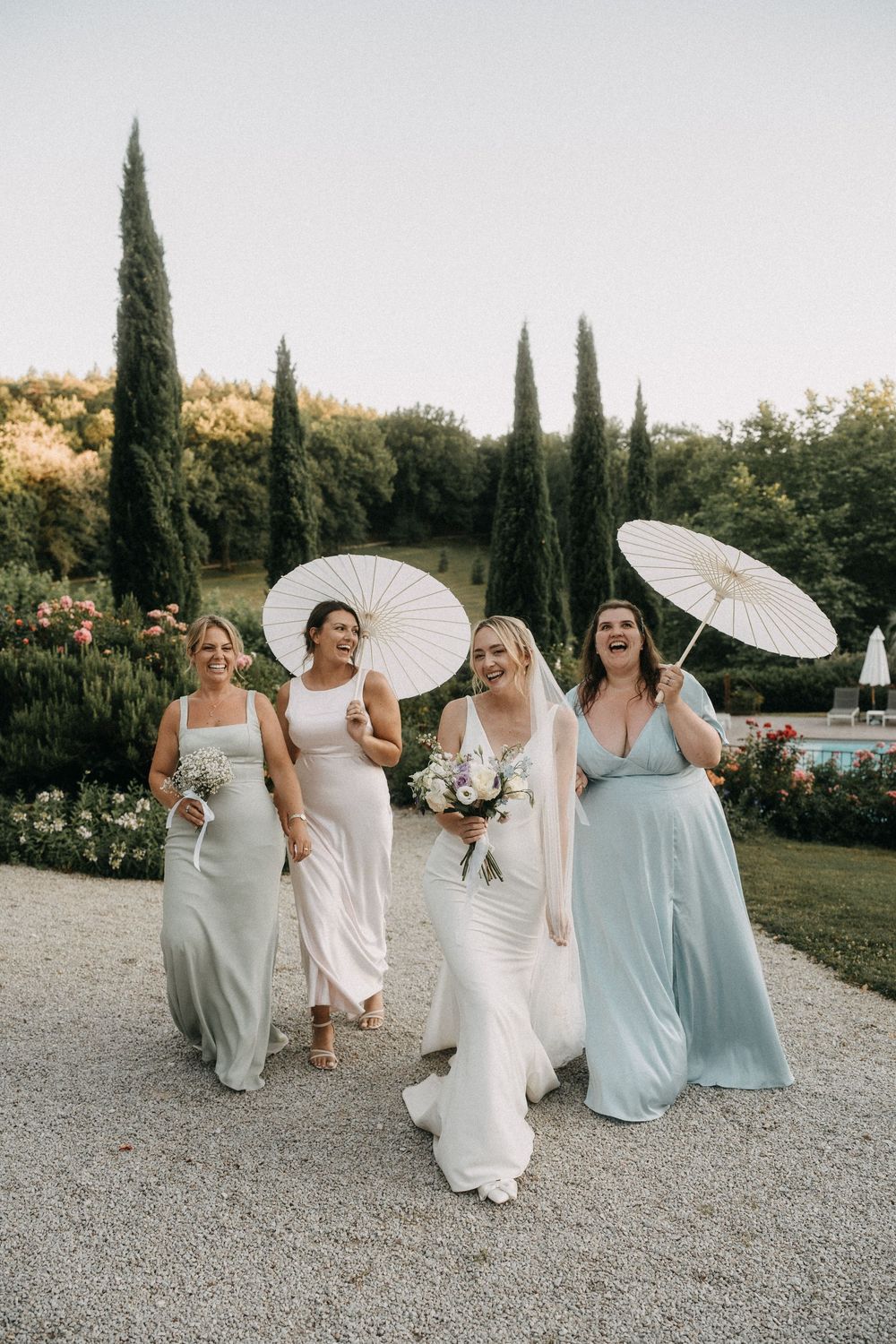 Four bridesmaids in flowing pastel dresses walk together holding white parasols at an outdoor garden wedding ceremony.