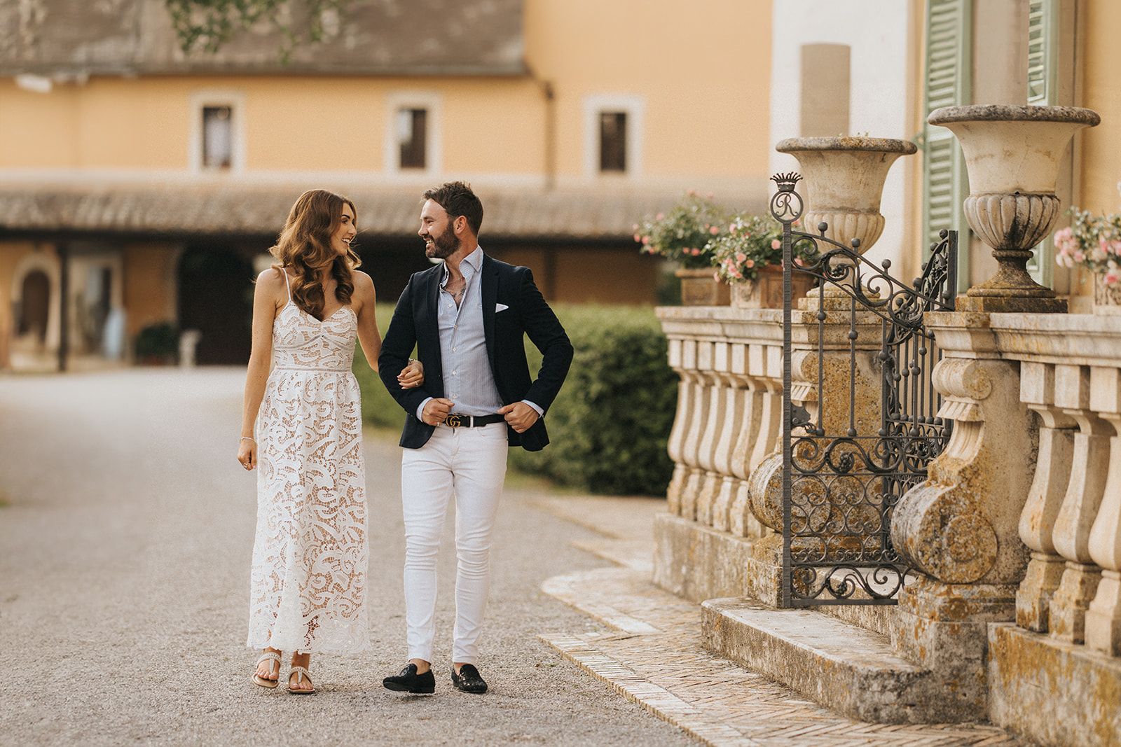 A couple in elegant attire walks hand in hand outside a luxurious Mediterranean villa on a sunny afternoon.