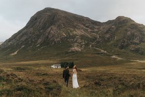 Two people in wedding attire stand together on a rugged hillside with a dramatic Scottish mountain peak in the background.
