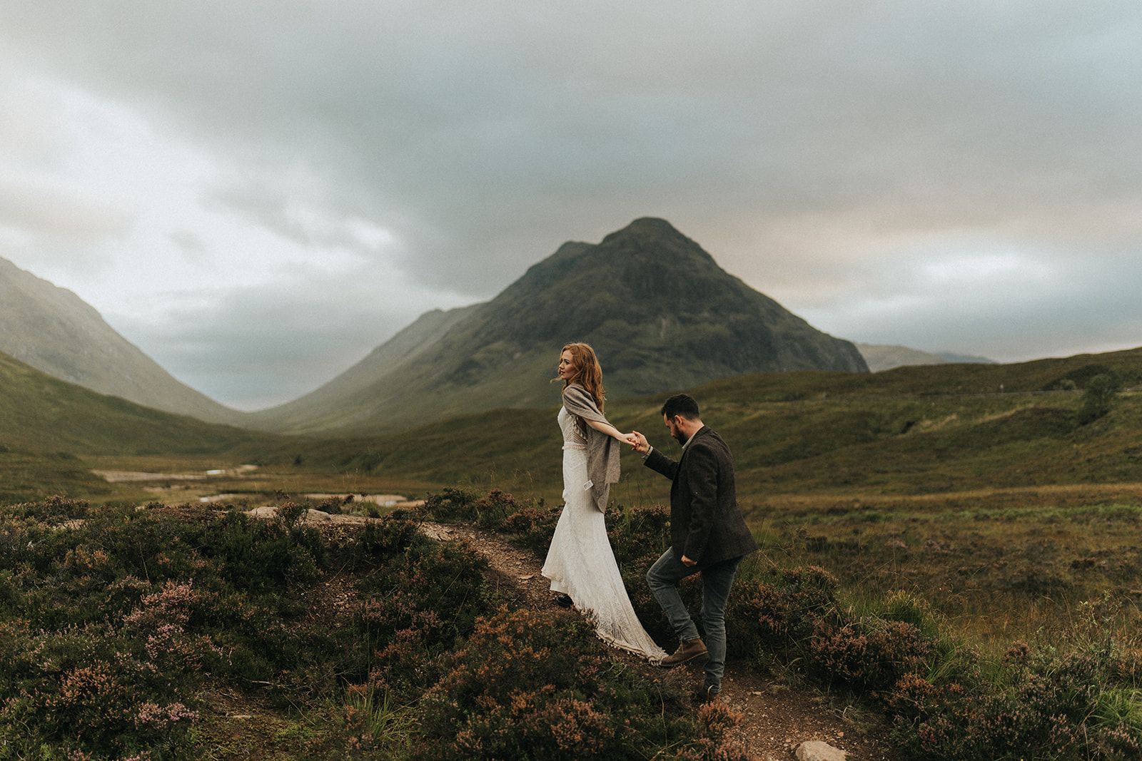 A couple walks through the dramatic Scottish Highlands landscape with moody clouds and rolling hills.