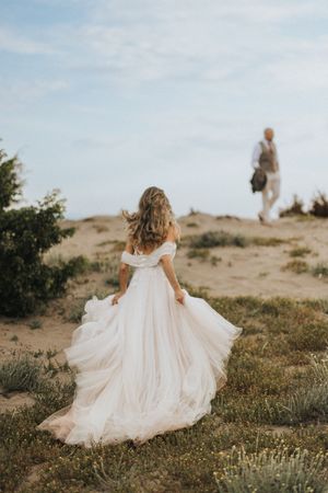 A person in a flowing white wedding dress runs up a sandy hill while another figure walks in the distance.