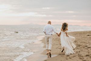 Two figures walk hand in hand along a sunset beach while wearing white formal attire and leaving footprints in the sand.