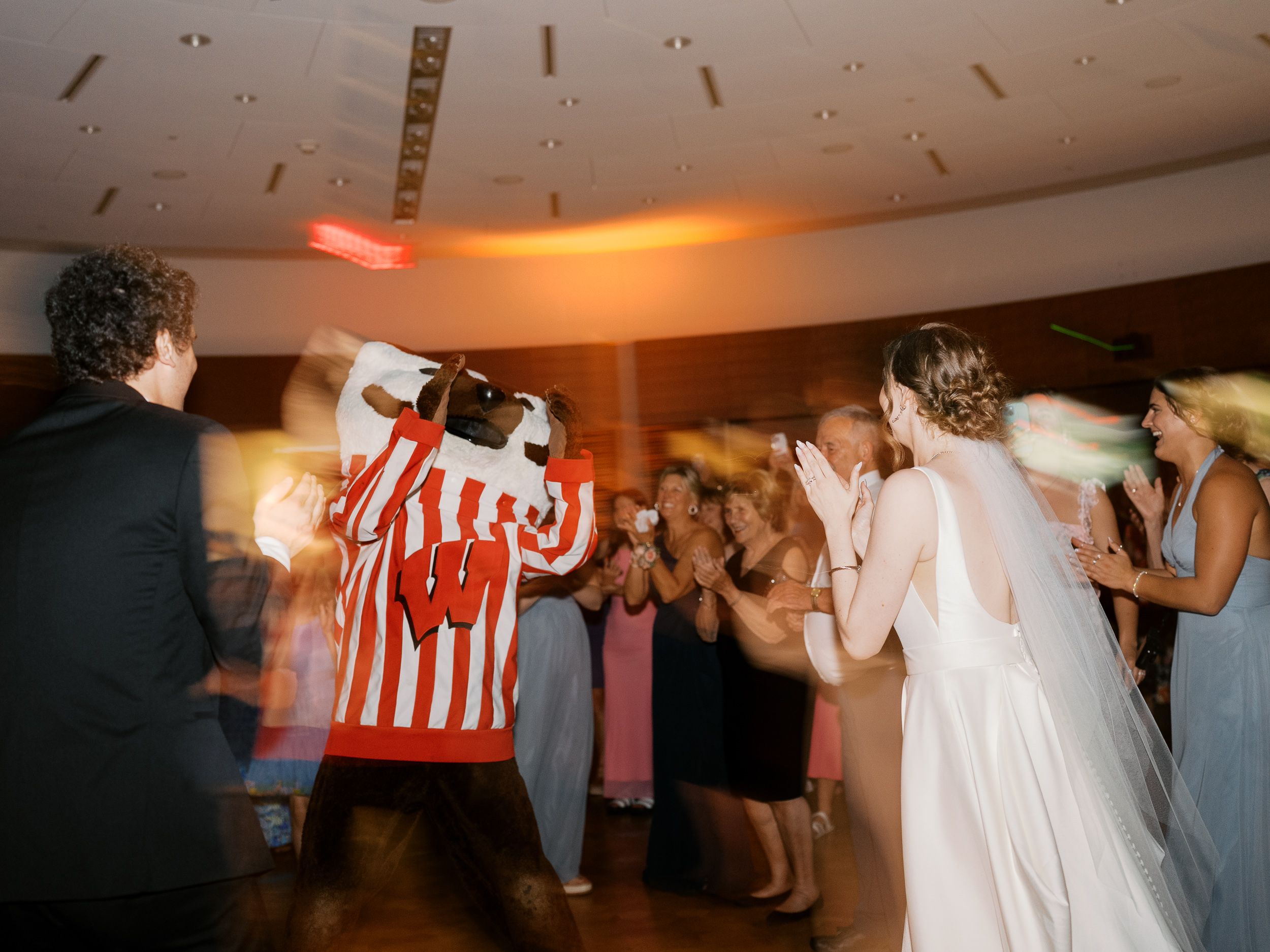 People dance in lines at an indoor social gathering with warm lighting and casual attire with Bucky Badger.