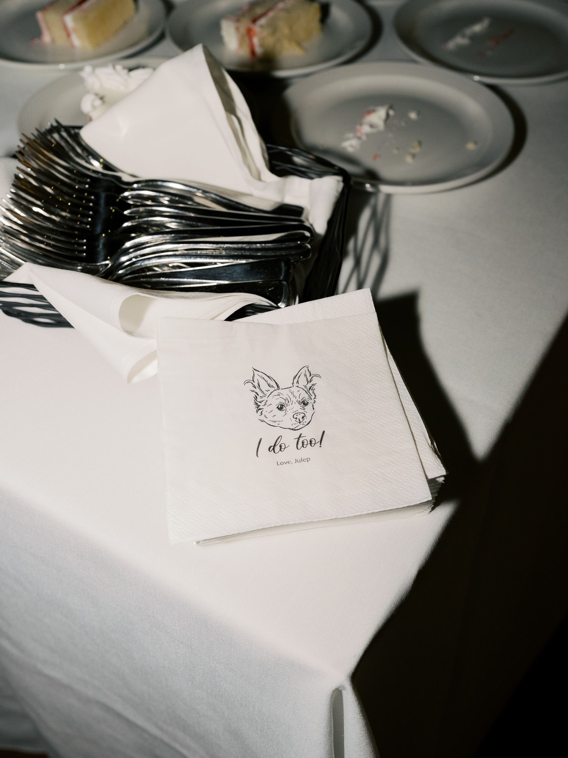 White wedding napkins with silverware and a simple dog logo design reading 'i do too!' arranged on a table setting.