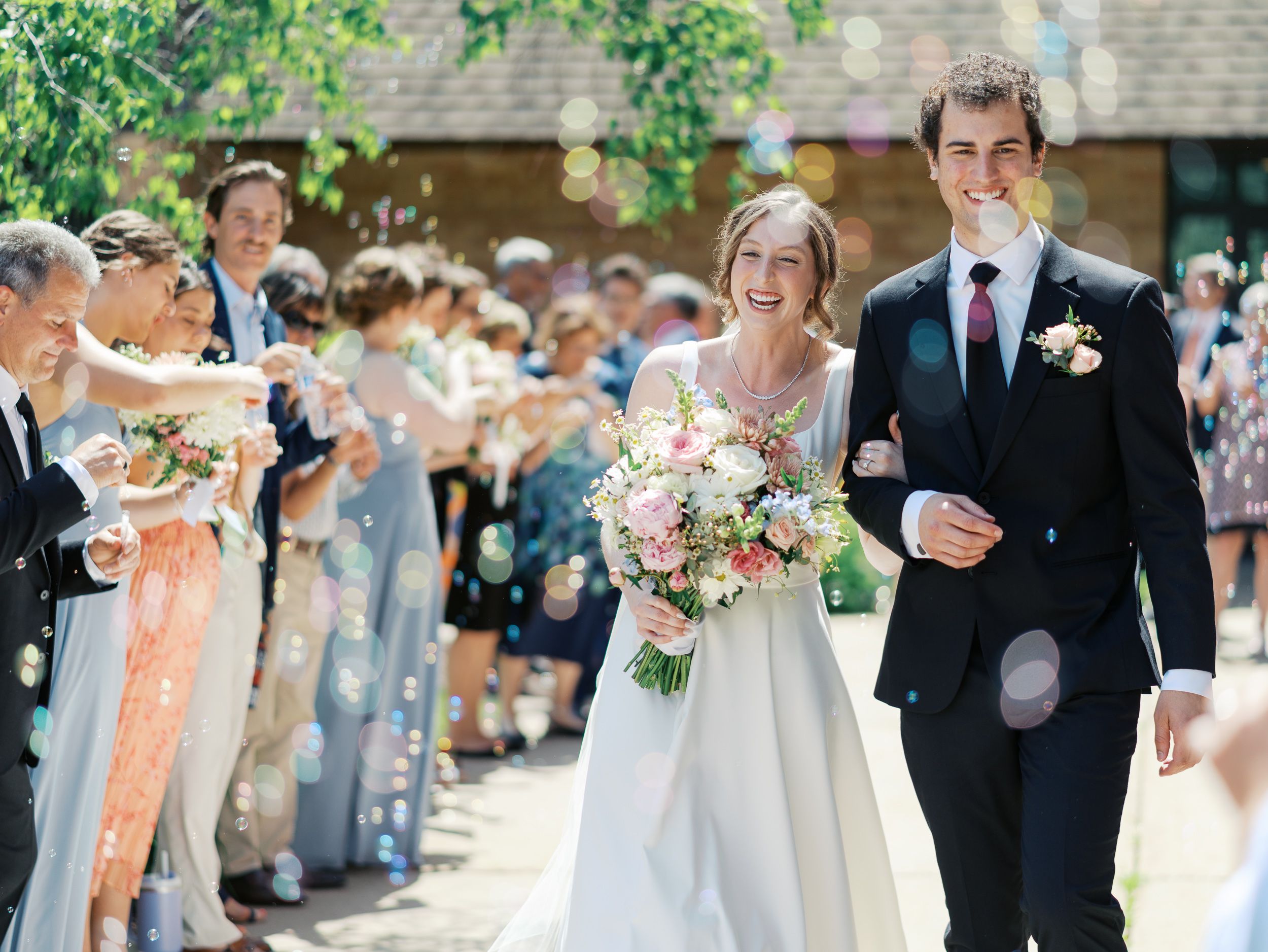 Wedding couple walks past guests throwing confetti in outdoor ceremony with bride holding pink and white bouquet.