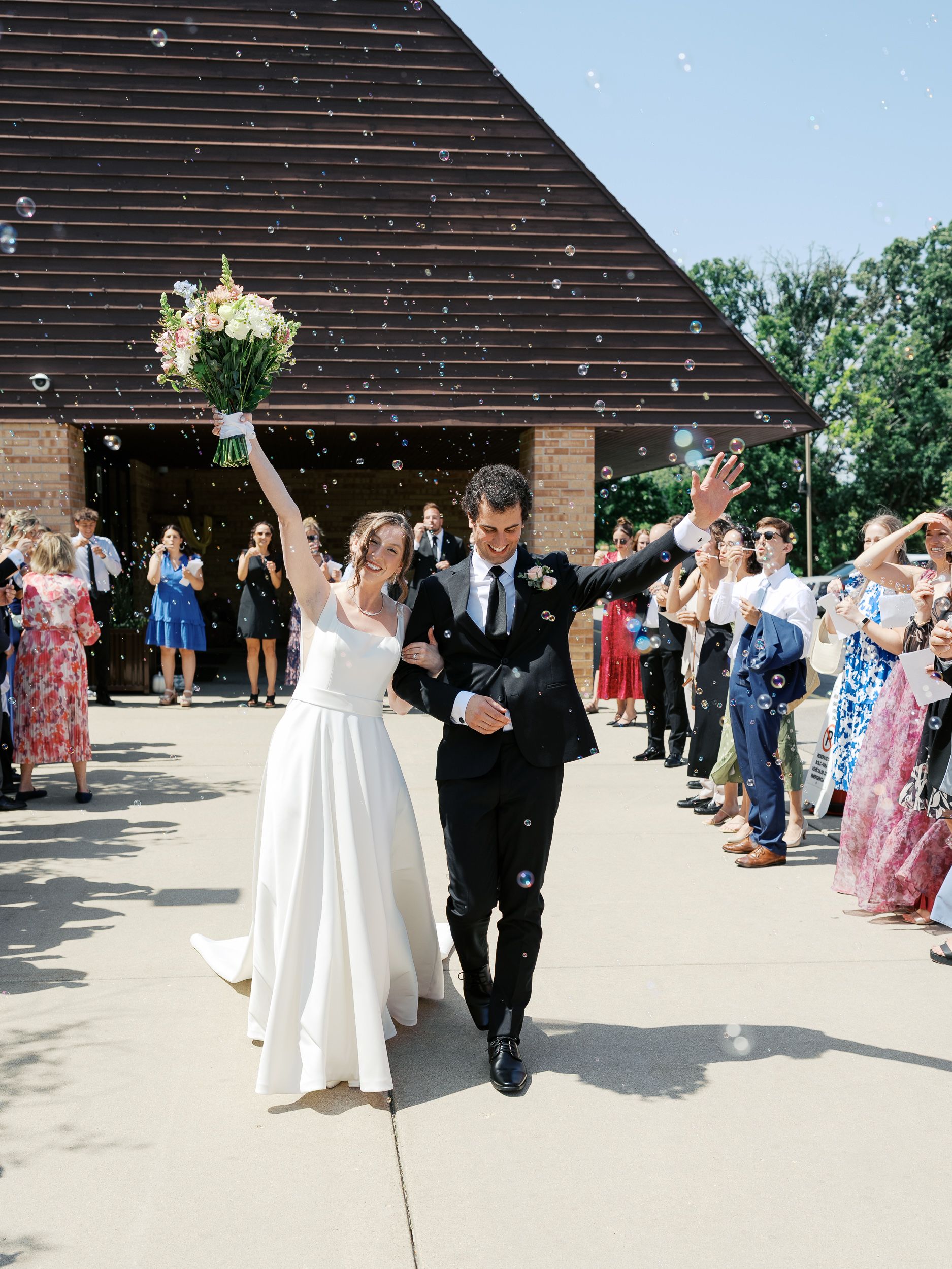 Wedding couple makes grand exit through confetti and bubbles as wedding guests celebrate outside a rustic venue.