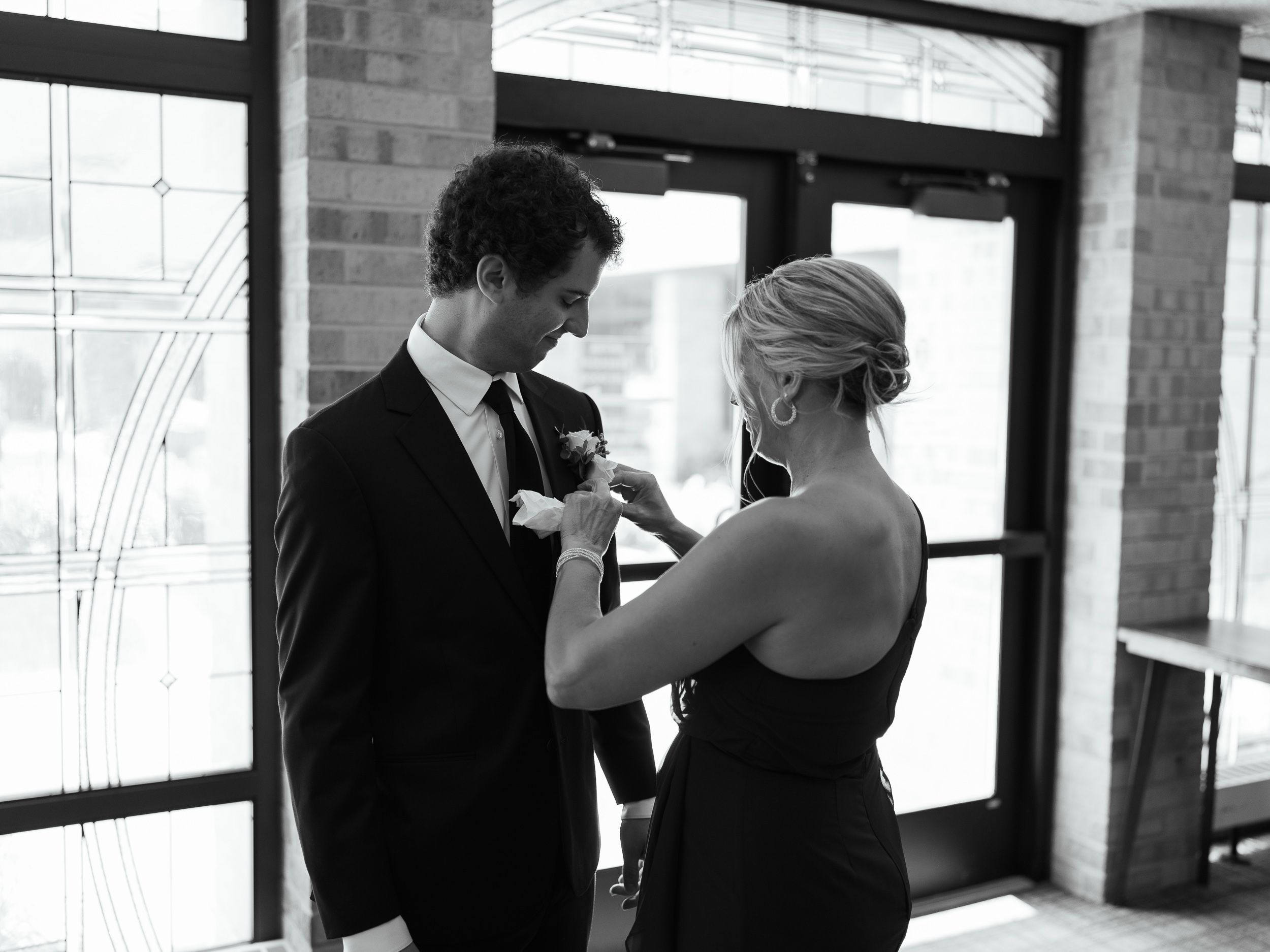 A tender moment at a formal event as someone adjusts the boutonniere on a suit jacket in black and white.