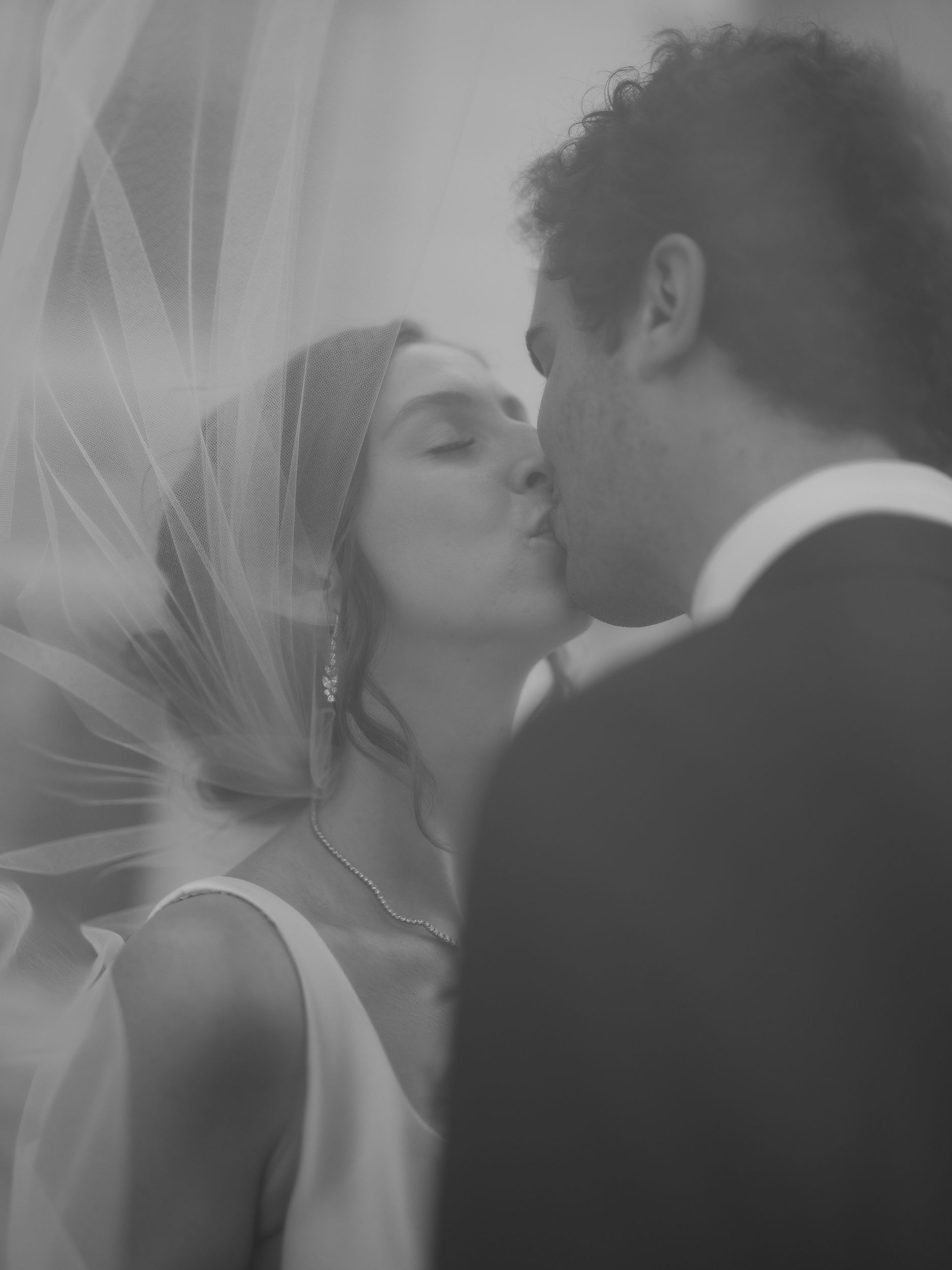 A romantic black and white wedding photo showing a couple sharing a tender kiss with a flowing veil in soft light.