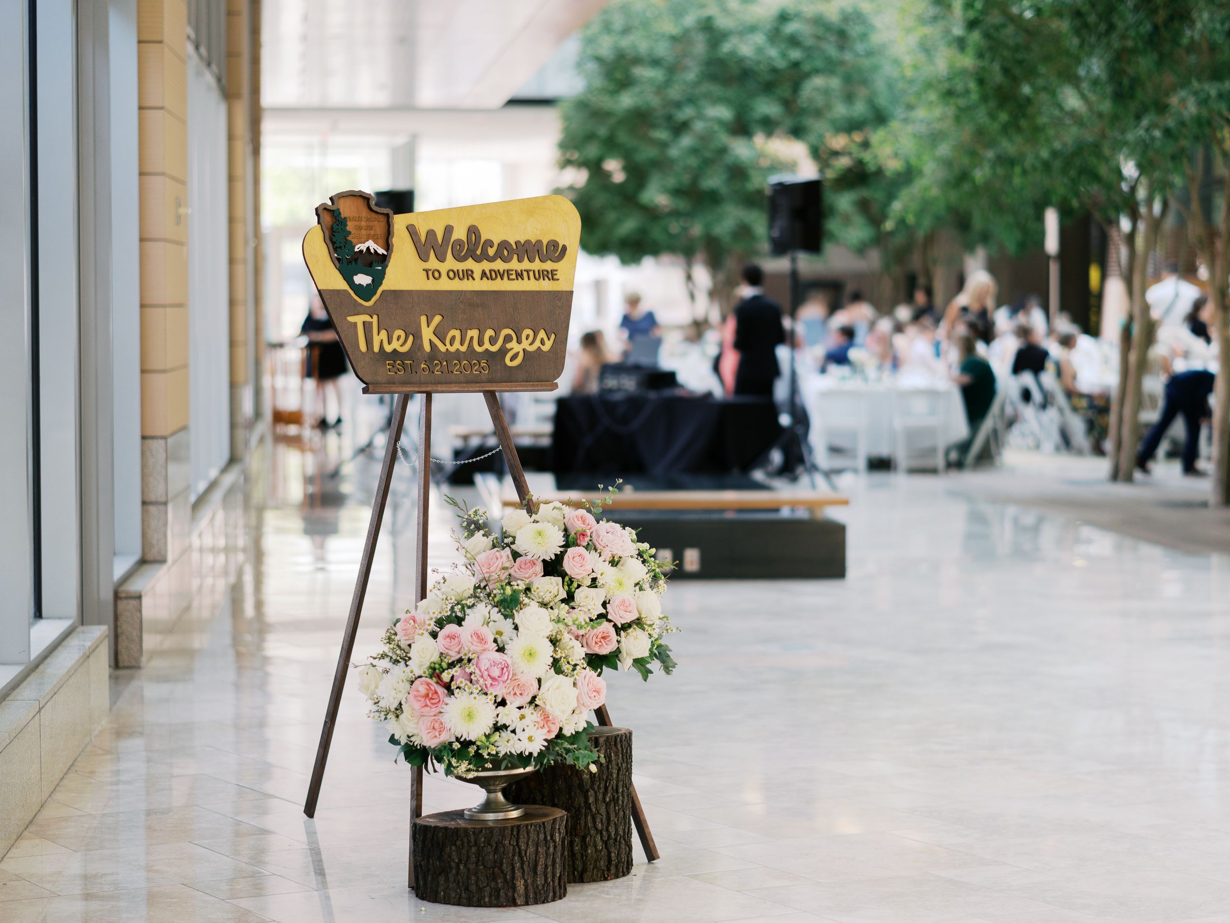 Welcome sign and floral arrangement on wood slices decorate a venue entrance with guests mingling in background.