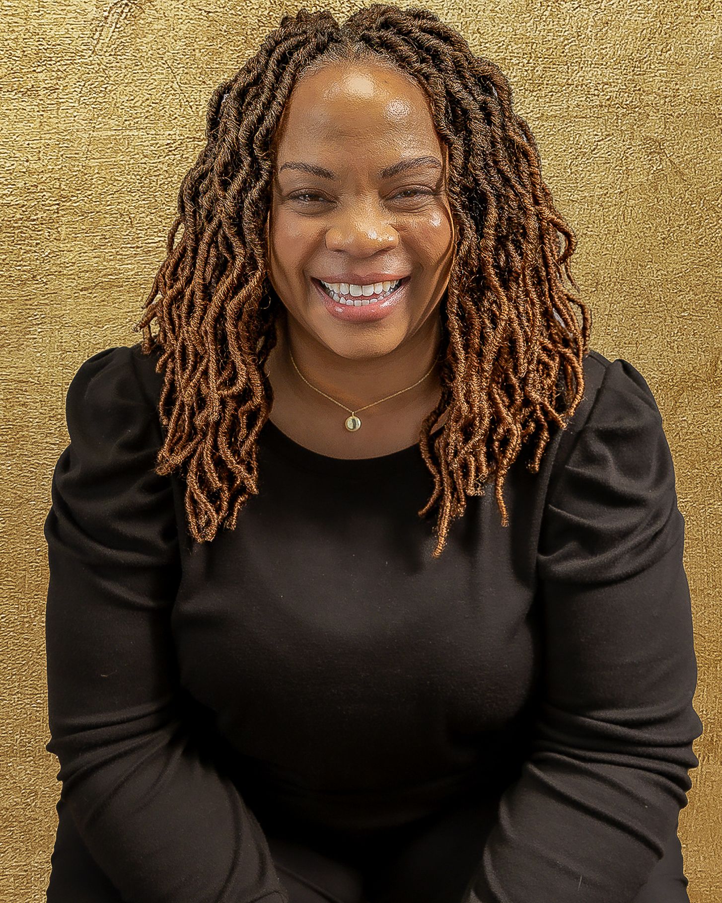 A person with copper-toned locs wearing a black sweater smiles warmly against a gold textured background.