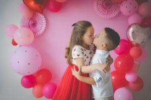A young brother and sister giving each other a little kiss framed by a Valentines balloon garland in pinks and reds.
