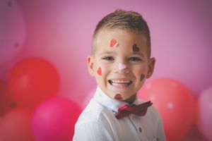 A boy with heart sickers all over his face smiling at the camera with a red bowtie and Valentine colour theme balloons in the background