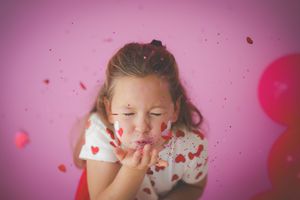 A girl blowing little paper hearts toward the camera on a pink backdrop