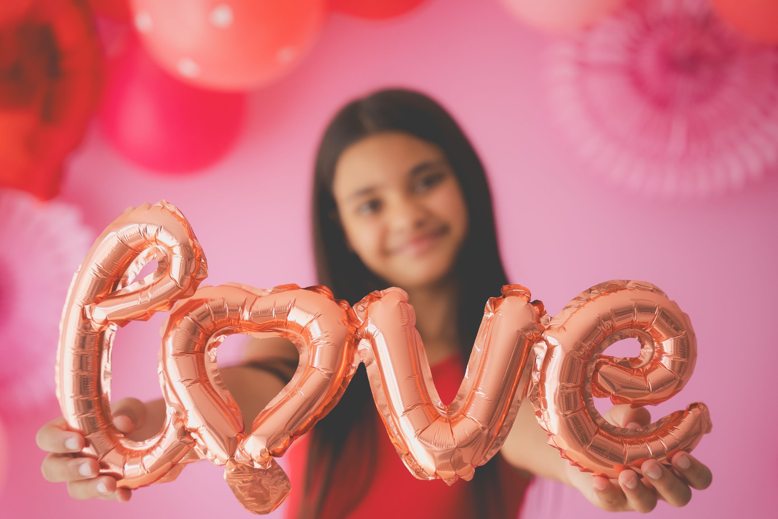 A girl holding a foil balloon showing the letters LOVE with pink and red valentines decoreations in the background