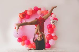 A Valentines background of pink and red balloons while a yound girl does a one handed handstand
