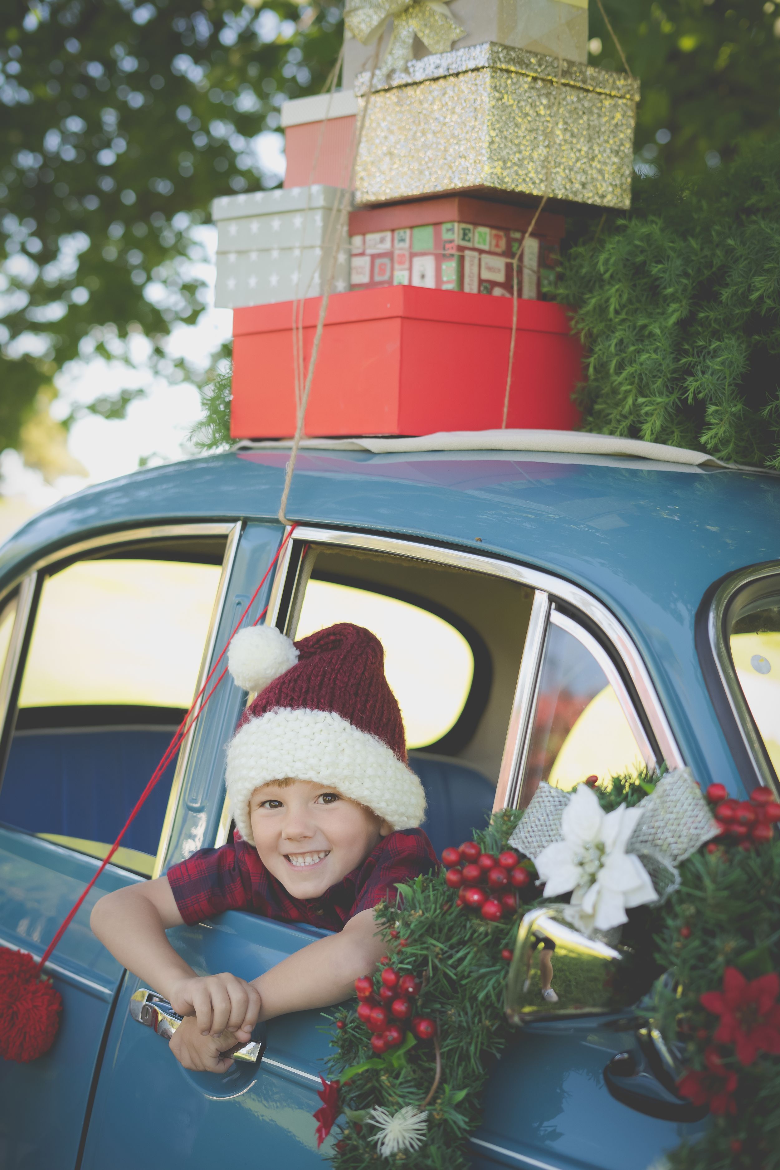 A blue vintage morris minor car in the background laden with Christmas gifts and a tree while a bou sits out the window wearing a hat