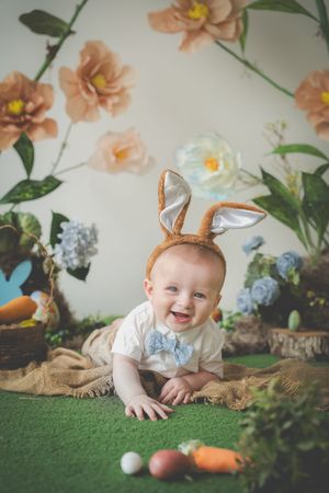 A happy toddler in a Easter themed setting of giant flowers, easter eggs and carrots. He is earing a little bowtie and easter bunny ears
