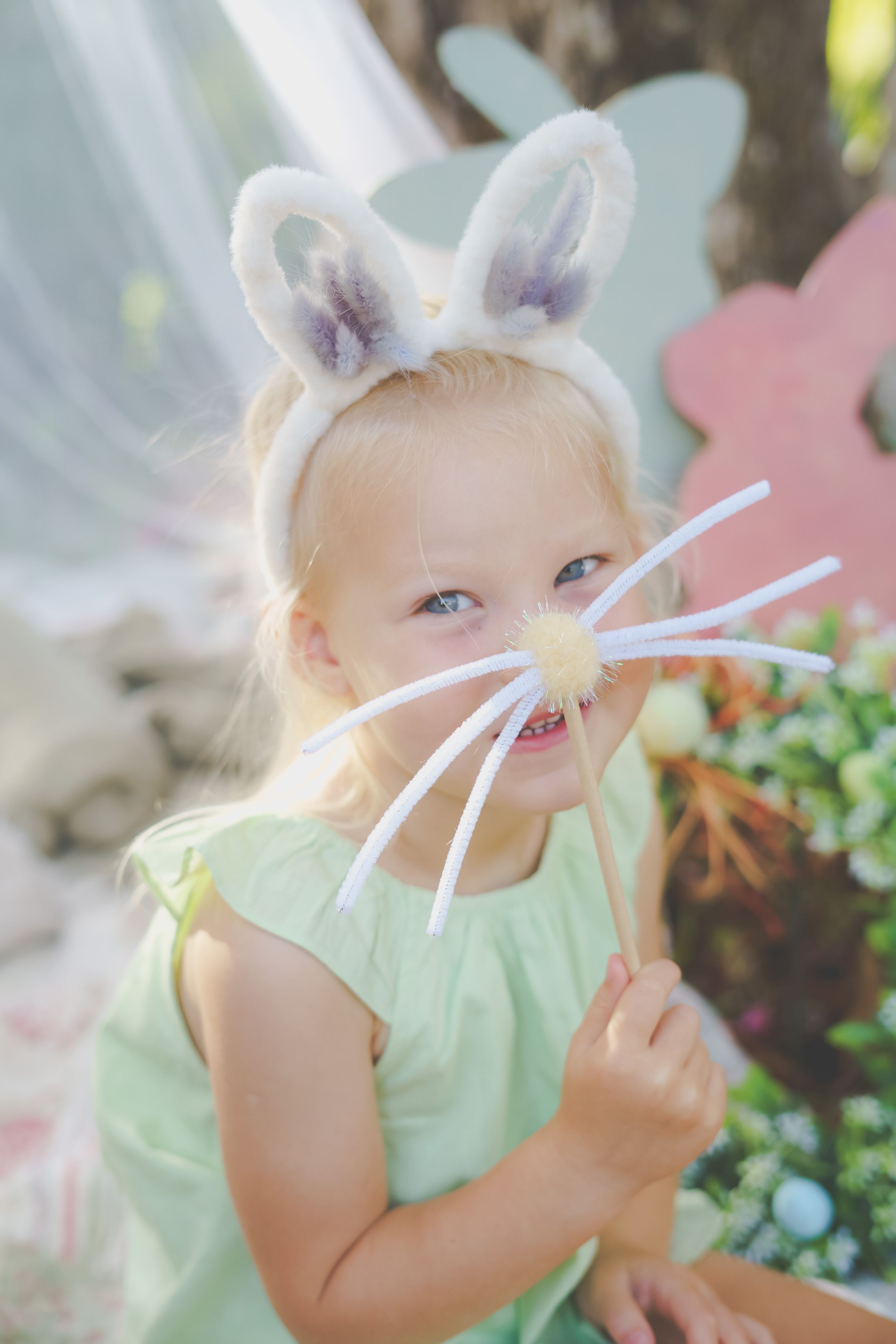 A young girl holding up a home made easter bunny ears and nose and whiskers smiling at the camera