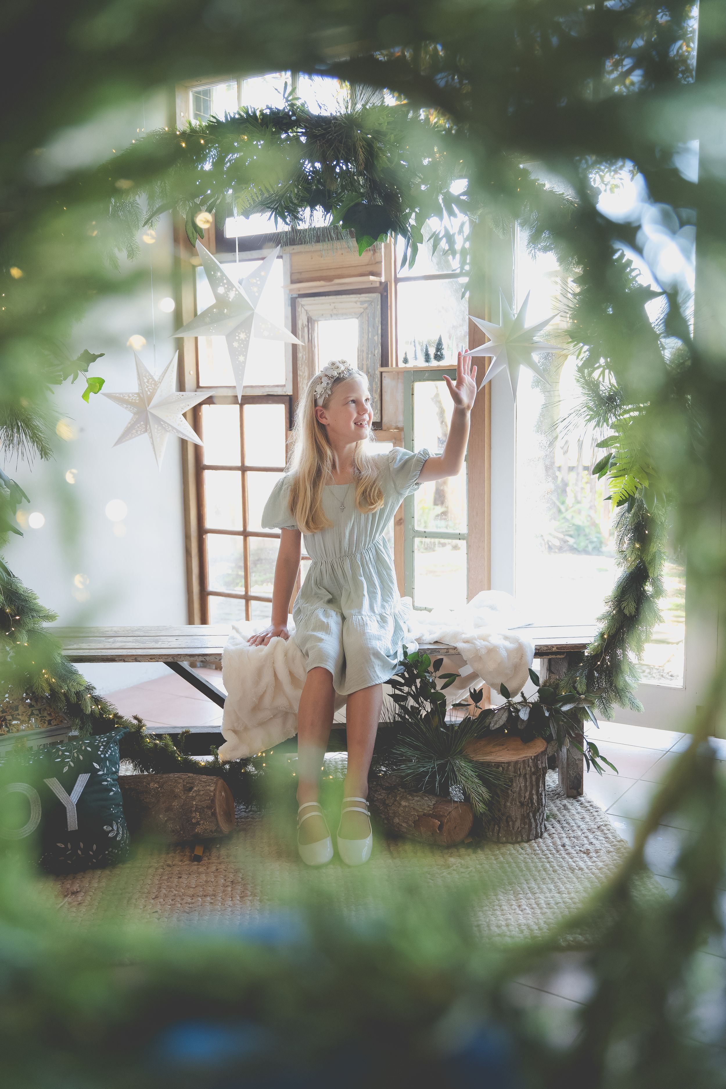 A Christmas scene of a girl reaching for a Christmas bauble surrounded by a big wreather with windows in the background