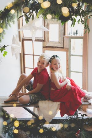 A Christmas scene of borther and sister sitting back to back on a wooden bench surrounded by Christmas lights and greenery