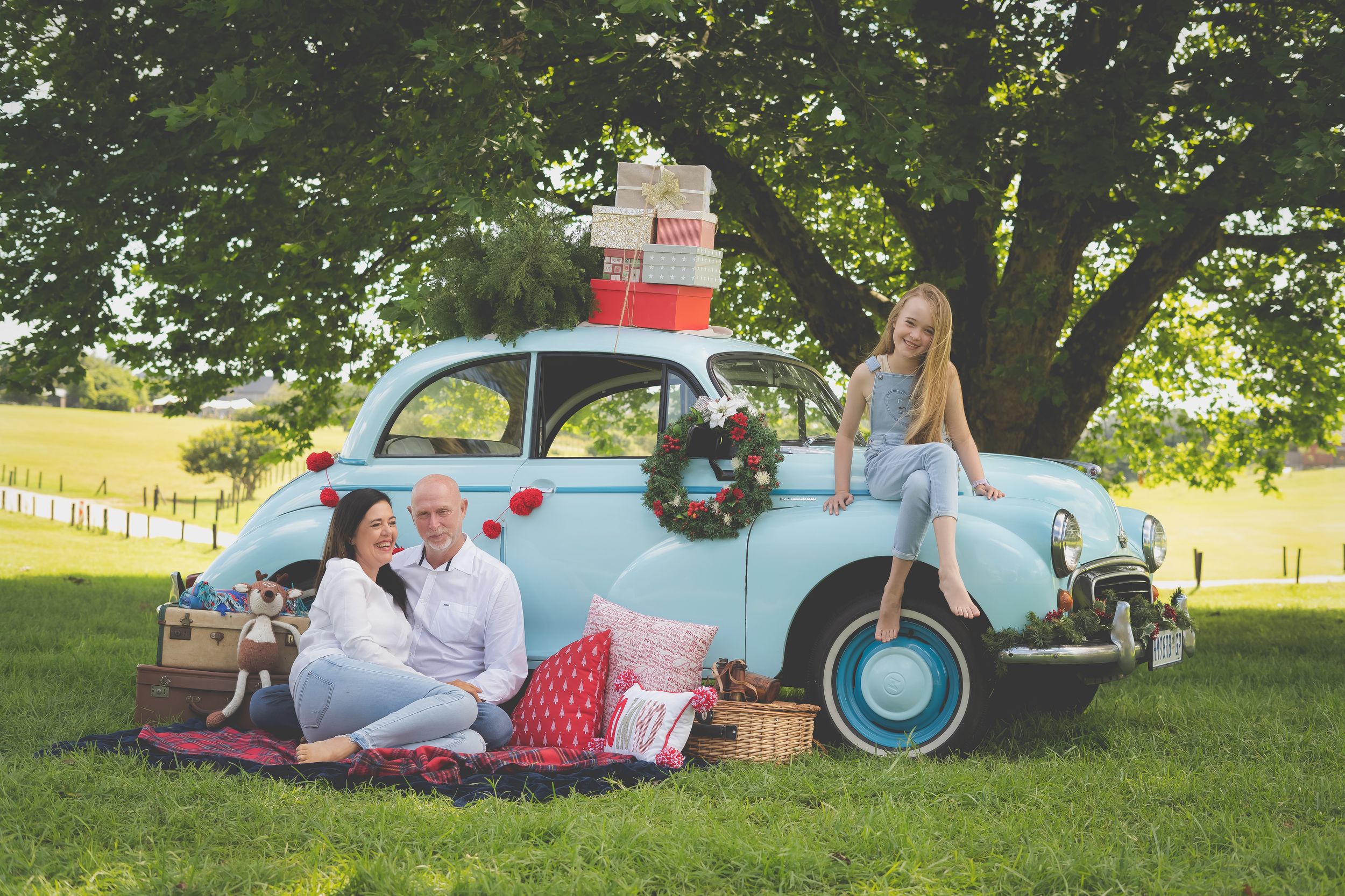 A blue vintage morris minor car in the background laden with Christmas gifts and a tree while a family poses with a picnic blanket