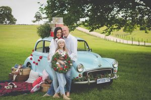 A blue vintage morris minor car in the background laden with Christmas gifts and a tree while a family poses on the bonet