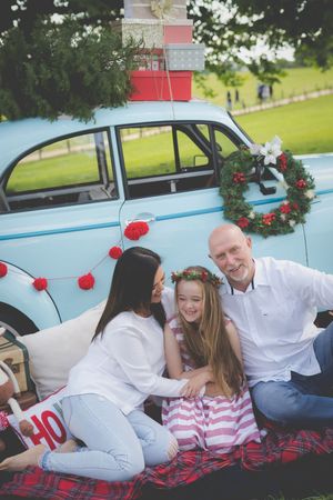 A blue vintage morris minor car in the background laden with Christmas gifts and a tree while a family laughts on a picnic blanket