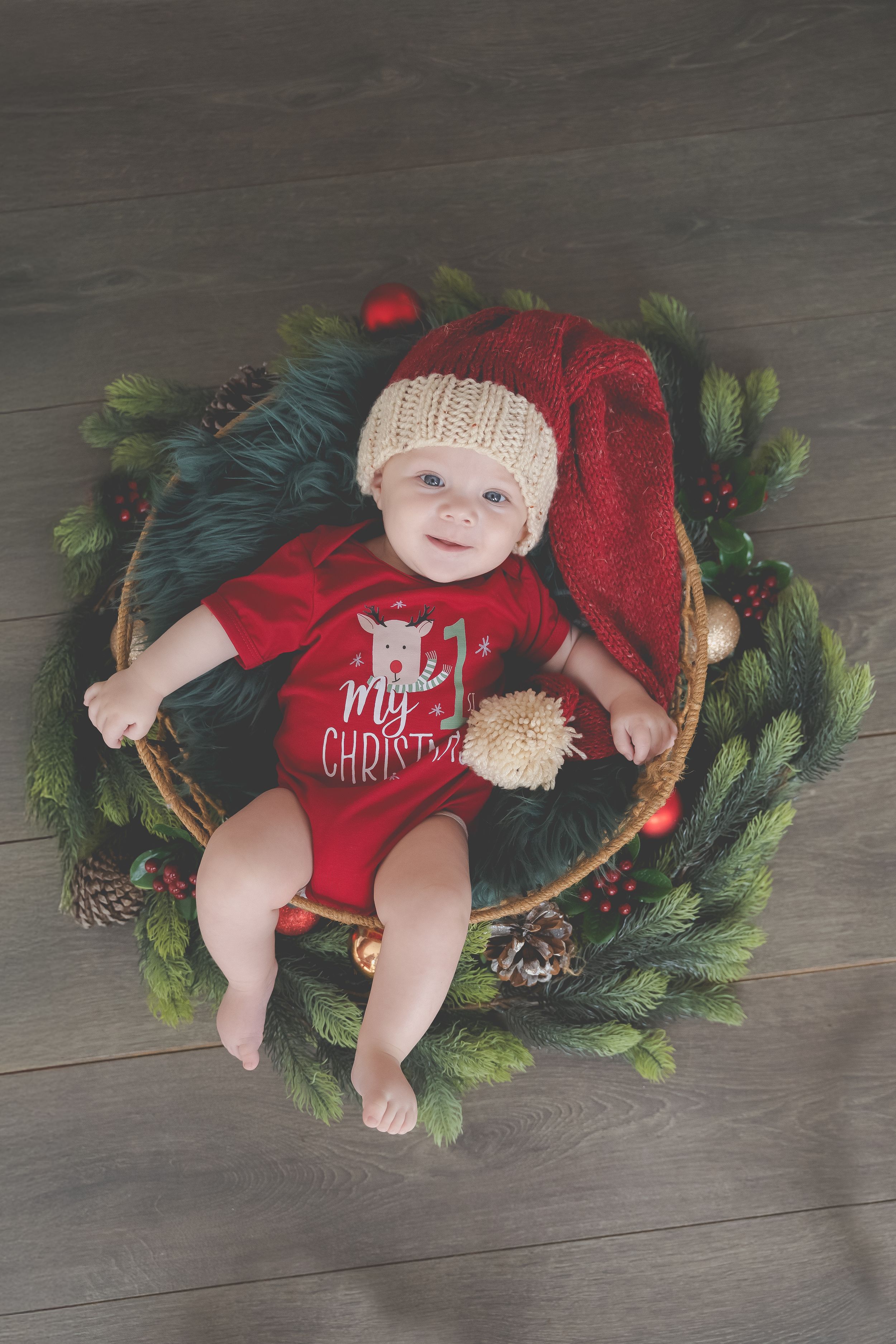 A baby dressed in a Christmas romper and hat, lying in a basket surrounded by a decorated wreath.
