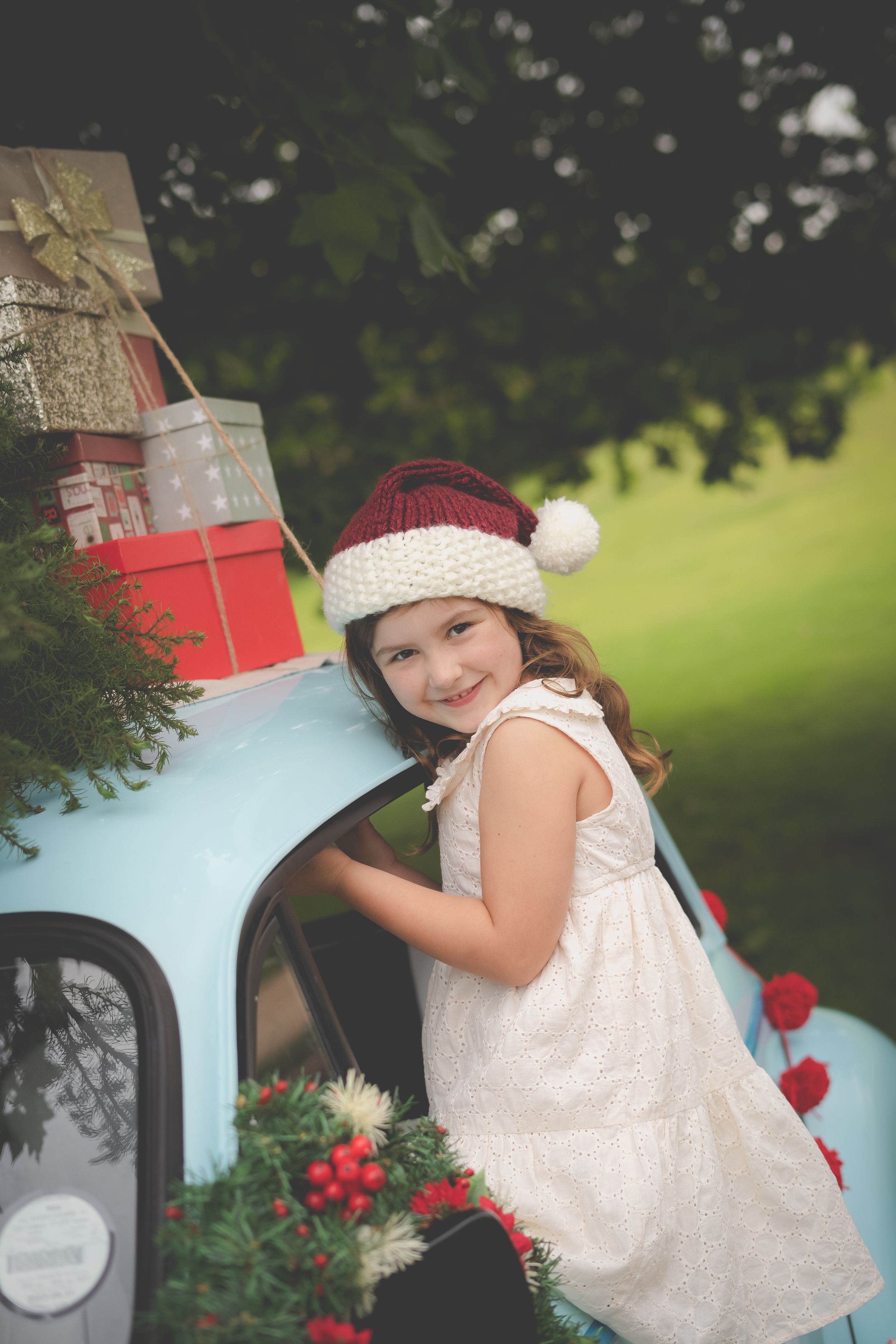 A blue vintage morris minor car in the background laden with Christmas gifts and a tree while a young girl sits out the window wearing a hat