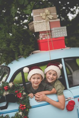A blue vintage morris minor car in the background laden with Christmas gifts and a tree while two brother stick their heads out the window
