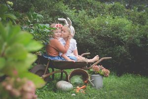 An Easter scene in the garden, brother and sister sit in an old wheelbarrow together, with bunny ears and decorations around.