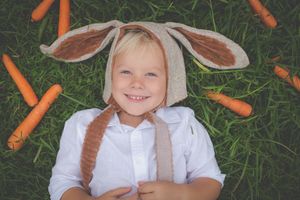 A boy smiling boy dressed in a bunny eared hat and lying on his back in the long grass surrounded by carrots