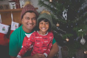 A young boy sitting on his dad's lap both dressed in Christmas theme cloting and smiling at the camera while tree lights the background