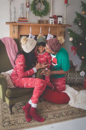 A Christmas living room scene with mom and dad kissing their son on the cheeks a fireplace, Christmas tree and decorations in the background