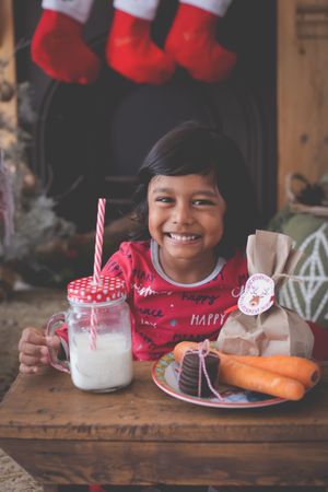 A young boy grinning at the camera with his milk and cookies for Santa and a fireplace and stockings in the background