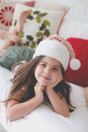 A beautiful little girl lying on a bed wearing a Christams had and Christmas scatter cushions in the background