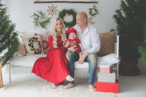 A family Christmas portrait in red and greens with wreaths and Christmas trees in the background. A smiling family on a cound in foreground